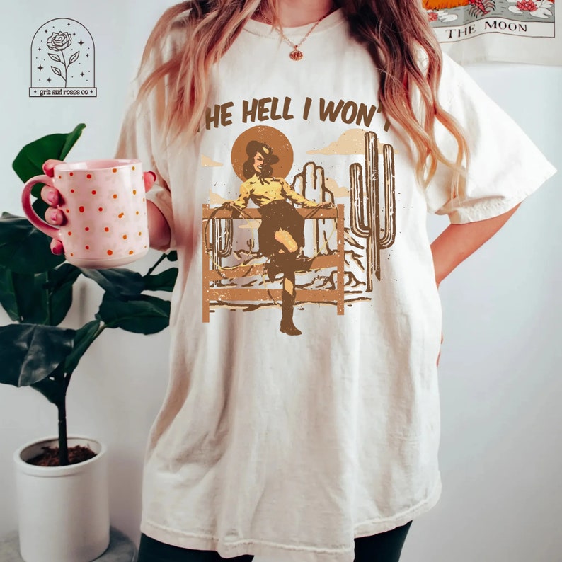 May include: A beige t-shirt with the text "The Hell I Won't" printed on it. The graphic features a woman in a cowboy hat and boots, standing on a fence with a lasso in her hand. The background is a desert scene with cacti.