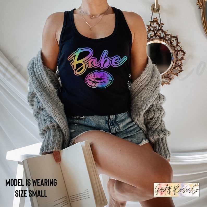 Puede incluir: Un top negro de tirantes con un gr&aacute;fico degradado arco&iacute;ris que dice "Babe" y una impresi&oacute;n de labios degradados arco&iacute;ris. La modelo lleva una talla peque&ntilde;a.