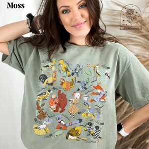 May include: A sage green t-shirt with a colorful graphic print of animals and books. The graphic features a variety of animals, including birds, a rooster, a bear, a rabbit, a frog, and a goose. The animals are all wearing clothes and holding books. The text "Moss" is printed in white at the top of the shirt.
