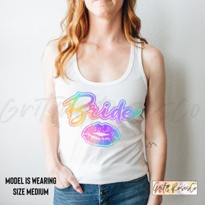 Puede incluir: Camiseta blanca de tirantes con el texto "Bride" en degradado arco&iacute;ris y una impresi&oacute;n de labios en degradado arco&iacute;ris.