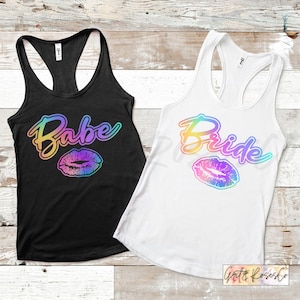 Puede incluir: Dos camisetas de tirantes con un dise&ntilde;o degradado arco&iacute;ris. Una camiseta de tirantes es negra con el texto "Babe" y una impresi&oacute;n de labios arco&iacute;ris. La otra camiseta de tirantes es blanca con el texto "Bride" y una impresi&oacute;n de labios arco&iacute;ris.