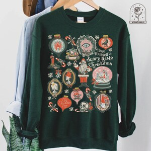 May include: A dark green sweatshirt with a festive design featuring a variety of spooky Christmas ornaments, including a skull, a bat, a mushroom, and a snowman. The text "Have Yourself a Scary Little Christmas" is printed on the front of the sweatshirt.