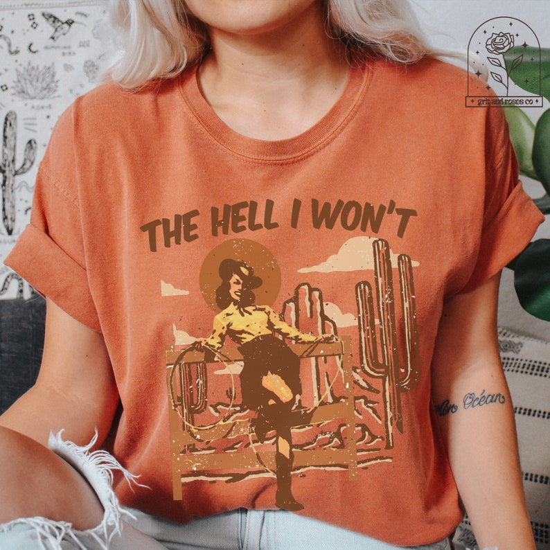 May include: A rust-colored t-shirt with a vintage graphic of a woman in a cowboy hat and a lasso. The text "THE HELL I WON'T" is printed above the graphic.