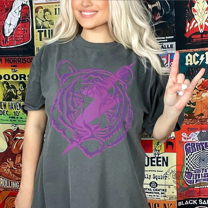 Pu&ograve; includere: Una maglietta grigia scura con una grafica di tigre viola e un design a forma di fulmine. Gli occhi della tigre sono viola.