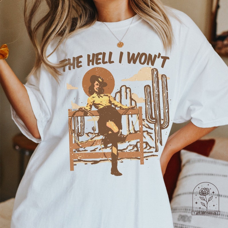 May include: White t-shirt with a graphic of a woman in a cowboy hat and boots, standing on a fence with a lasso. The text "THE HELL I WON'T" is printed above the graphic.