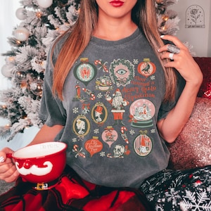 May include: A gray t-shirt with a vintage Christmas design featuring a skull, a skeleton, a mushroom, a Santa Claus, and a snow globe. The text on the shirt reads "Have yourself a scary little Christmas".