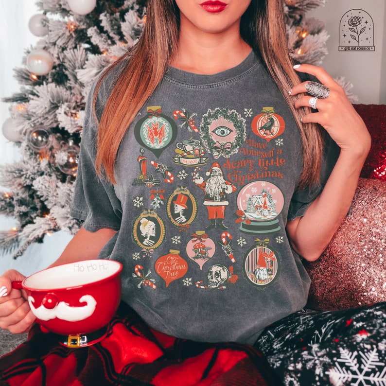 May include: A gray t-shirt with a vintage Christmas design featuring a skull, a skeleton, a mushroom, a Santa Claus, and a snow globe. The text on the shirt reads "Have yourself a scary little Christmas".
