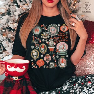 May include: A black t-shirt with a festive, spooky Christmas design. The shirt features a variety of illustrations, including a skull, a Christmas tree, a gnome, a snow globe, and a Santa Claus. The text on the shirt reads "Have Yourself a Scary Little Christmas".