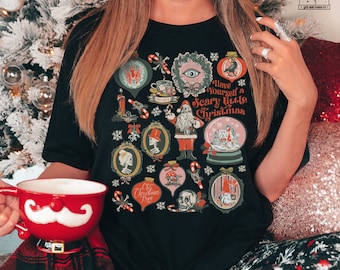 May include: A black t-shirt with a festive, spooky Christmas design. The shirt features a variety of illustrations, including a skull, a Christmas tree, a gnome, a snow globe, and a Santa Claus. The text on the shirt reads "Have Yourself a Scary Little Christmas".
