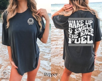 Puede incluir: Una camiseta negra de gran tamaño con una cara sonriente a cuadros en la parte delantera y el texto "Wake up & smell the race fuel" en la parte posterior. El texto está en una fuente retro con un borde a cuadros.
