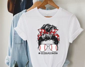 May include: White t-shirt with a graphic of a woman with a messy bun, red and white polka dot headband, and baseball-themed sunglasses. The text "#TEEBALLMOM" is printed below the graphic.