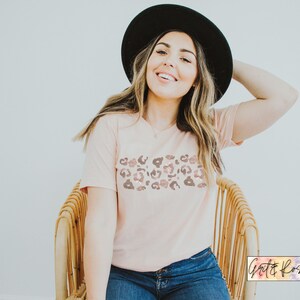 May include: A woman wearing a pink t-shirt with a brown and pink leopard print design. She is wearing a black hat and blue jeans. The t-shirt has a v-neck and short sleeves.