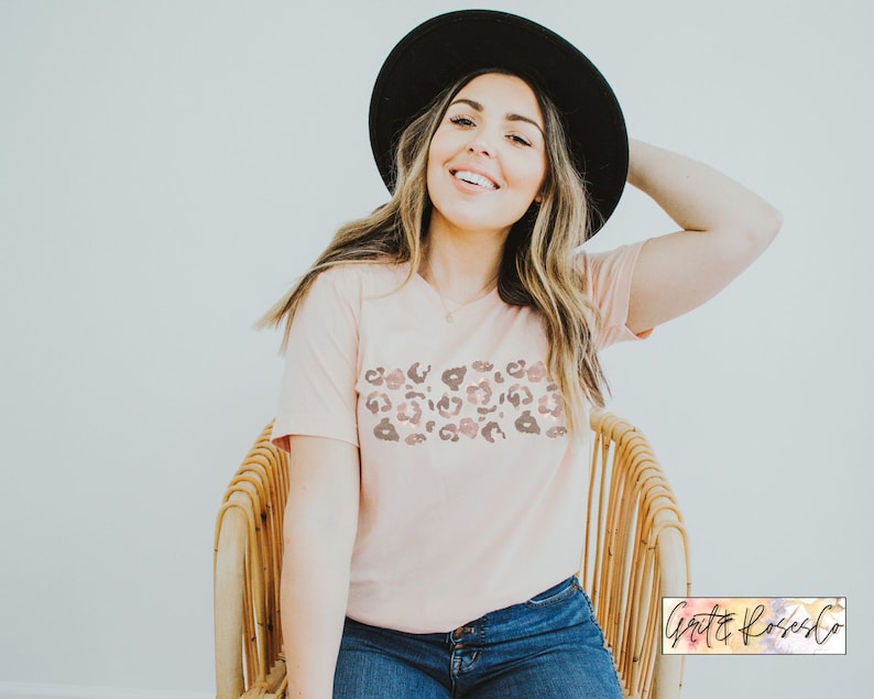May include: A woman wearing a pink t-shirt with a brown and pink leopard print design. She is wearing a black hat and blue jeans. The t-shirt has a v-neck and short sleeves.