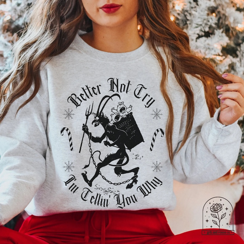 May include: Light gray sweatshirt with a black graphic of a devil carrying a basket with babies, candy canes, and snowflakes. The text reads "Better Not Try, I'm Tellin' You Why" in a gothic font. A festive, holiday-themed design.