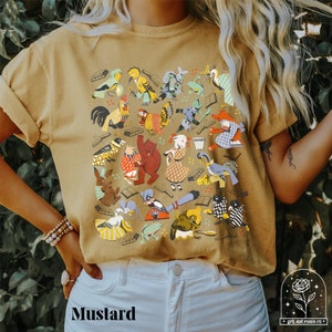May include: A mustard yellow t-shirt with a colorful graphic print of various animals and objects. The graphic includes a rooster, a pig, a frog, a bird, a rabbit, and a book. The shirt has a short sleeve and a crew neck.