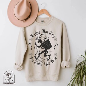 May include: Beige sweatshirt with a black graphic of a devil carrying a sack, pitchfork, and chain. The text reads "Better Not Try, I'm Tellin' You Why." Includes snowflakes and candy cane accents.