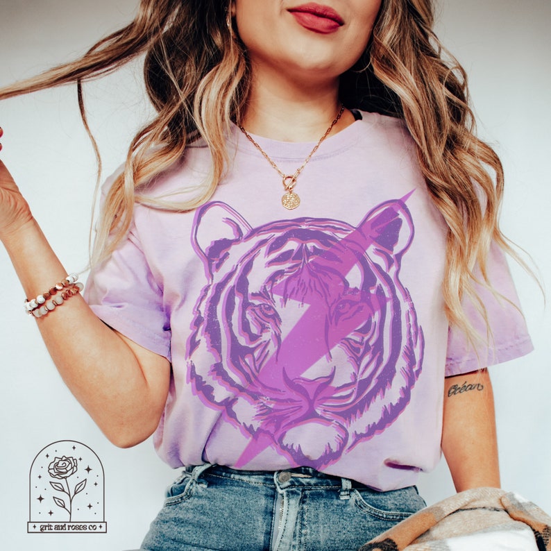 Pu&ograve; includere: Una maglietta tie-dye viola con una grafica di tigre viola e bianca e un design a forma di fulmine. La maglietta ha il testo "Gobern" scritto sulla manica.