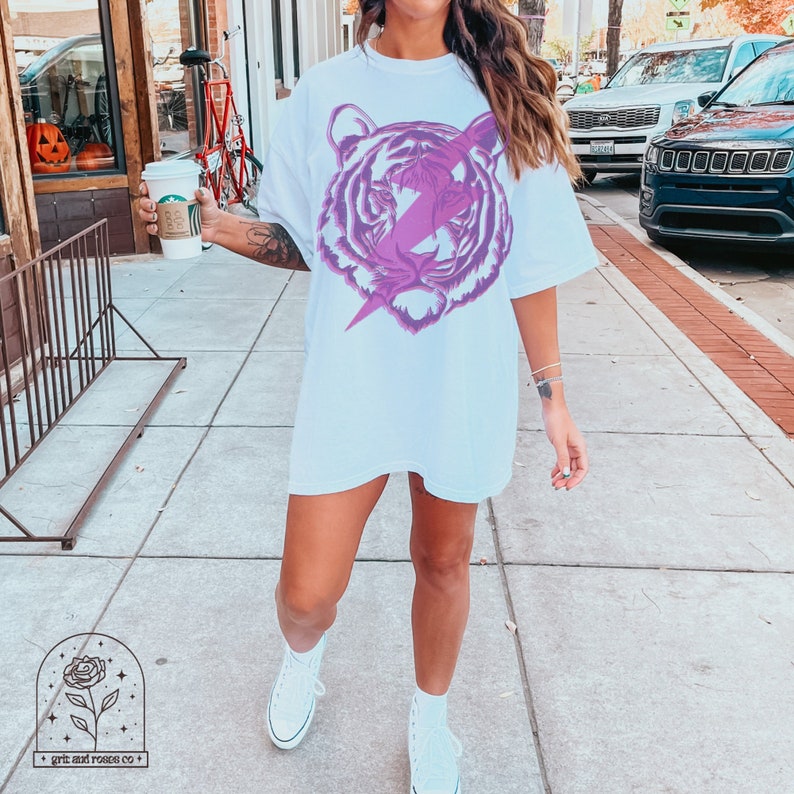 Pu&ograve; includere: Maglietta bianca oversize con una grafica viola di una testa di tigre con un fulmine che la attraversa.