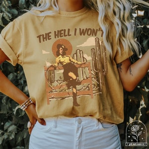 May include: A mustard yellow t-shirt with a vintage graphic of a woman in a cowboy hat riding a horse. The text "THE HELL I WON'T" is printed on the shirt.