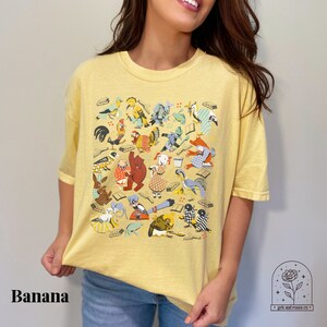 May include: A yellow t-shirt with a colorful illustration of animals and objects. The illustration includes a rooster, a bear, a frog, a bird, a rabbit, and a fox. The text "Banana" is visible at the bottom of the image.