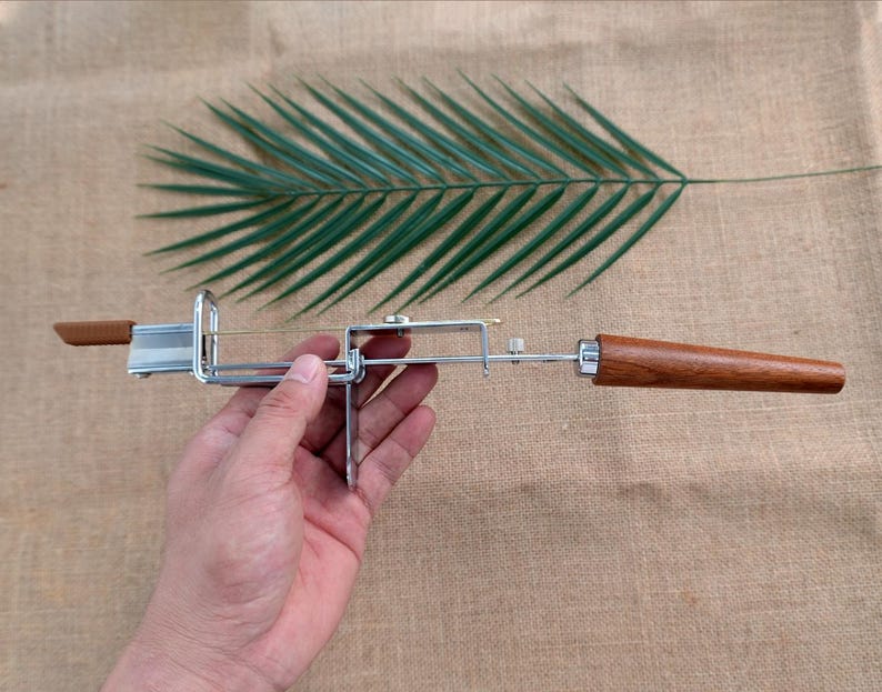 Op de afbeelding: Een kaassnijder met een roestvrijstalen frame en houten handvatten. De snijder heeft een draadmes en is ontworpen voor het snijden van kaas. De houten handvatten hebben een warme bruine kleur.