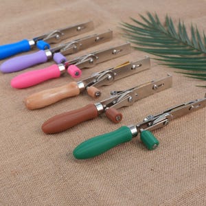 May include: A collection of metal and wood tools with colorful handles. The handles are blue, purple, pink, light brown, dark brown, and green. The tools are arranged in a row on a textured surface.