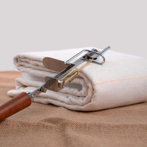 Op de afbeelding: Een metalen en houten gereedschap met een mes rust op een gevouwen witte doek met oranje strepen. Het gereedschap heeft een houten handvat en een zilveren metalen frame. De doek is gevouwen op een bruin oppervlak.