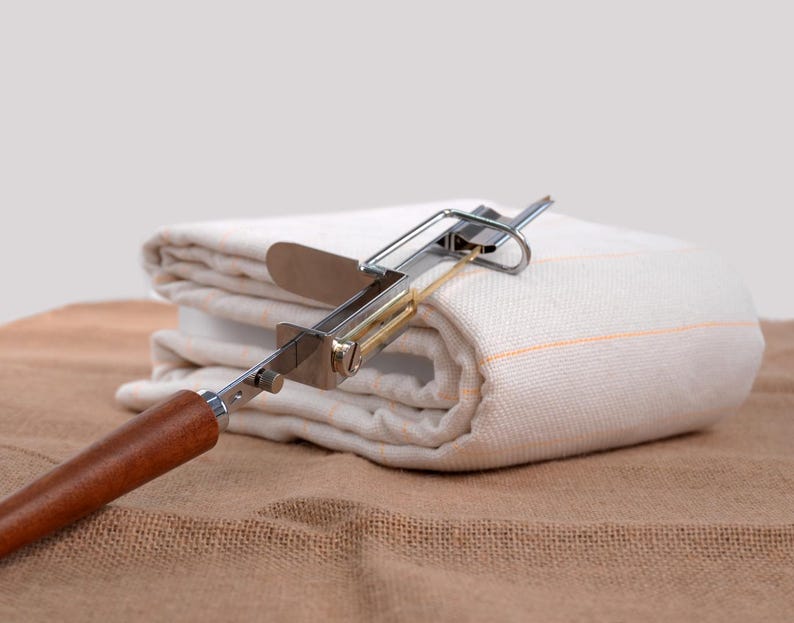 Op de afbeelding: Een metalen en houten gereedschap met een mes rust op een gevouwen witte doek met oranje strepen. Het gereedschap heeft een houten handvat en een zilveren metalen frame. De doek is gevouwen op een bruin oppervlak.