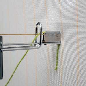 Op de afbeelding: Close-up van een metalen tapijthaakgereedschap met een zilveren afwerking, bevestigd aan een wit canvas met verticale oranje strepen. Een gele draad is over het gereedschap gespannen en een groen garen is bevestigd.