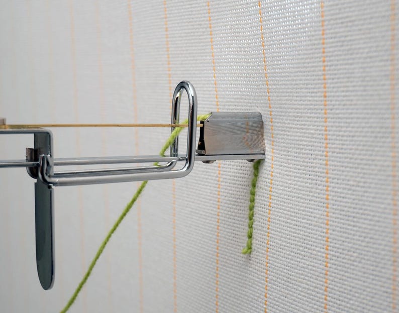 Op de afbeelding: Close-up van een metalen tapijthaakgereedschap met een zilveren afwerking, bevestigd aan een wit canvas met verticale oranje strepen. Een gele draad is over het gereedschap gespannen en een groen garen is bevestigd.