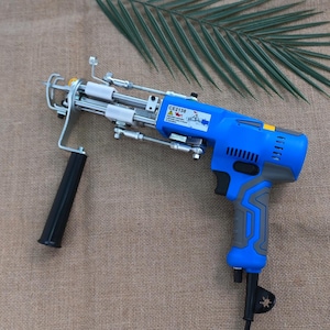Pode incluir: Uma pistola de tufos azul e cinza com uma alça preta e uma estrutura de metal prateado. A ferramenta tem uma etiqueta CE2138 e foi projetada para criar tapetes e outras artes têxteis. O fundo é uma superfície marrom texturizada.