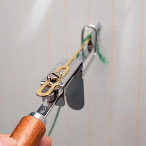 Op de afbeelding: Een zilver-gouden tapijttuftpistool met een houten handvat wordt gebruikt. De tool is gepositioneerd tegen een witte stof met groen garen dat erdoorheen wordt gevoerd. De tool is ontworpen voor het maken van handgemaakte tapijten en textielkunst.