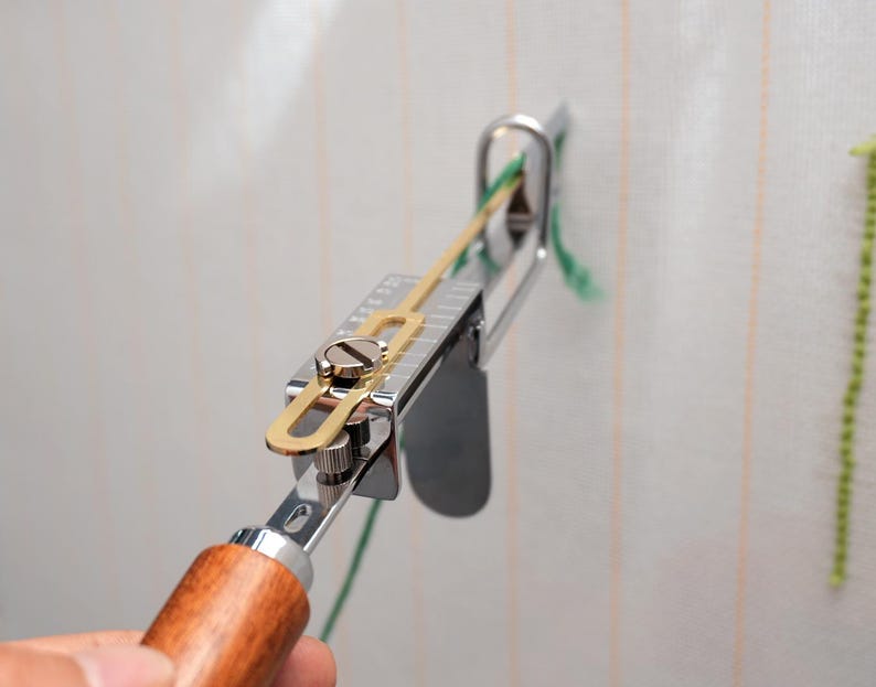 Op de afbeelding: Een zilver-gouden tapijttuftpistool met een houten handvat wordt gebruikt. De tool is gepositioneerd tegen een witte stof met groen garen dat erdoorheen wordt gevoerd. De tool is ontworpen voor het maken van handgemaakte tapijten en textielkunst.
