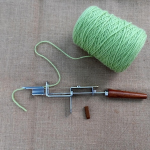 Op de afbeelding: Een lichtgroene bol garen, een metalen tapijt haakgereedschap en een klein houten stuk. De draad is verbonden met het gereedschap. Het gereedschap heeft een houten handvat. De items liggen op een beige stoffen oppervlak.
