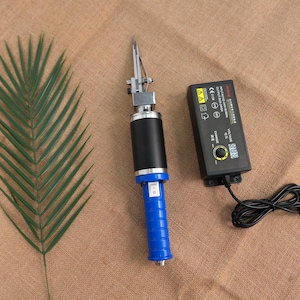 Può includere: Una pistola per ciuffi elettrica con un'impugnatura nera e blu, una testa in metallo argentato e un alimentatore nero. Una foglia di palma verde è sul lato sinistro dell'immagine.