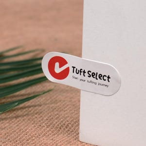 Op de afbeelding: Een ovale sticker met de tekst "Tuft Select" en de slogan "Start your tufting journey". De sticker heeft een rode cirkel met een wit vinkje. De sticker is op een wit oppervlak geplakt.