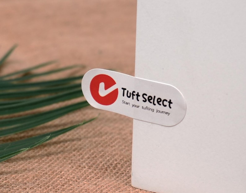 Op de afbeelding: Een ovale sticker met de tekst "Tuft Select" en de slogan "Start your tufting journey". De sticker heeft een rode cirkel met een wit vinkje. De sticker is op een wit oppervlak geplakt.