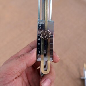 Op de afbeelding: Een zilverkleurig metalen gereedschap met een schuifschaal, met metingen van 7 tot 50. Het gereedschap heeft een messingkleurige component en een ronde instelknop. Het gereedschap wordt in een hand gehouden.