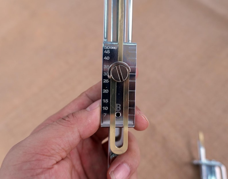Op de afbeelding: Een zilverkleurig metalen gereedschap met een schuifschaal, met metingen van 7 tot 50. Het gereedschap heeft een messingkleurige component en een ronde instelknop. Het gereedschap wordt in een hand gehouden.