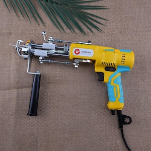 Può includere: Una pistola per ciuffi Tuft Select gialla e blu con manico e cavo neri. L'utensile ha una testa metallica con un ago e vari componenti meccanici. Lo sfondo è un tessuto marrone.