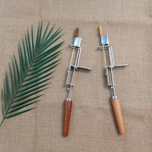 Può includere: Due utensili in metallo e legno con finitura argentata e manici in legno. Gli utensili hanno un telaio metallico rettangolare e un piccolo pezzo di legno nella parte superiore. Una foglia di palma verde è sul lato sinistro dell'immagine.