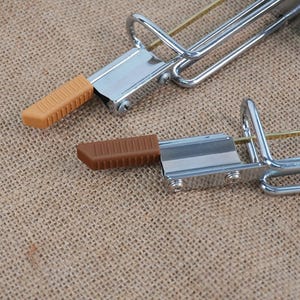 Op de afbeelding: Twee metalen klemmen met bruine en bruin rubberen handgrepen. De klemmen hebben een zilveren afwerking en een gebogen handvat. De rubberen handgrepen zijn rechthoekig met een geribbelde textuur. De achtergrond is een bruin, getextureerd weefsel.