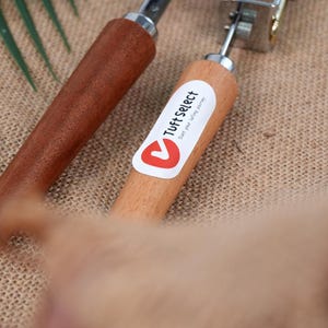 Op de afbeelding: Twee tapijttufts gereedschappen met houten handvatten. E&eacute;n handvat is donkerbruin van kleur, en de andere is lichtbruin met een witte sticker met de tekst "Tuft Select". De gereedschappen hebben metalen componenten.