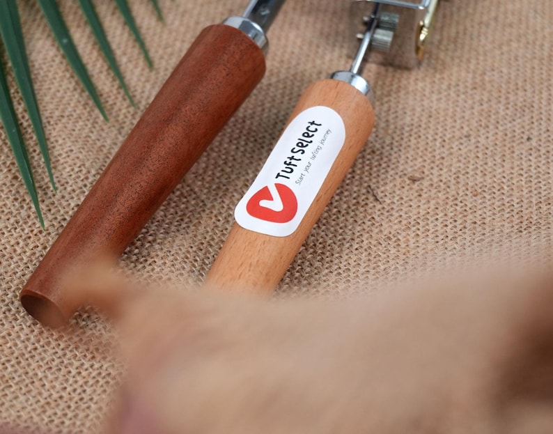 Op de afbeelding: Twee tapijttufts gereedschappen met houten handvatten. E&eacute;n handvat is donkerbruin van kleur, en de andere is lichtbruin met een witte sticker met de tekst "Tuft Select". De gereedschappen hebben metalen componenten.