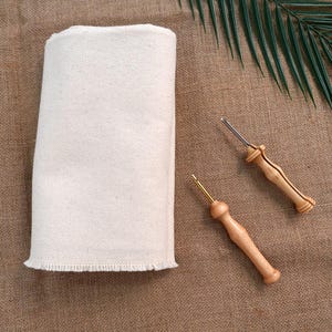 Op de afbeelding: Een opgerolde, crèmekleurige canvas stoffen rol met een gerafelde rand, naast twee houten punch needle tools. De tools hebben lichtbruine houten handvatten en metalen naalden. De achtergrond is een getextureerde, bruine stof.