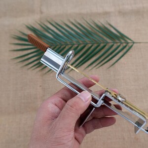 Op de afbeelding: Een metalen pastamessen met een bruin handvat en een zilveren frame. De snijder heeft een dun, goudkleurig mes en is ontworpen om pasta te snijden. De achtergrond heeft een groen blad en een bruin oppervlak.