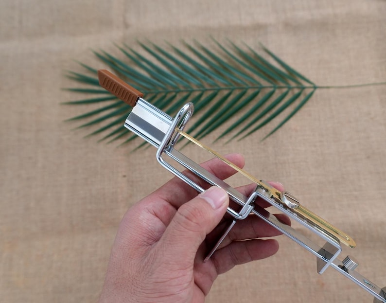 Op de afbeelding: Een metalen pastamessen met een bruin handvat en een zilveren frame. De snijder heeft een dun, goudkleurig mes en is ontworpen om pasta te snijden. De achtergrond heeft een groen blad en een bruin oppervlak.