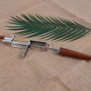 Op de afbeelding: Een zilverkleurige metalen sigarenknipper met een houten handvat. De knipper heeft een rechthoekig lichaam en een scharnierend mes. Een groen palmblad ligt op een bruin, getextureerd oppervlak.