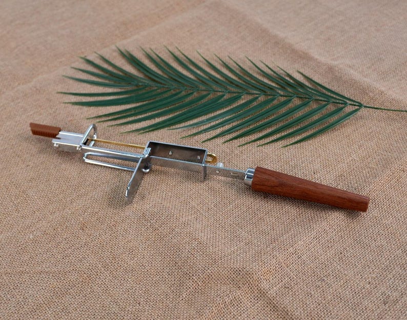 Op de afbeelding: Een zilverkleurige metalen sigarenknipper met een houten handvat. De knipper heeft een rechthoekig lichaam en een scharnierend mes. Een groen palmblad ligt op een bruin, getextureerd oppervlak.