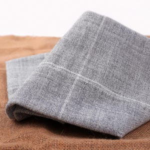 以下が含まれることがあります： 白いステッチのディテールが施された、2 つの折り畳まれた灰色のリネンナプキン。 ナプキンはテクスチャード加工された生地で作られており、茶色のジュートの表面に置かれています。 ナプキンはニュートラルカラーです。